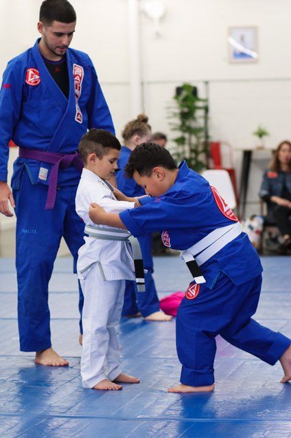 Two boys practicing Jiu Jitsu for beginners while a purple belt coach observes.